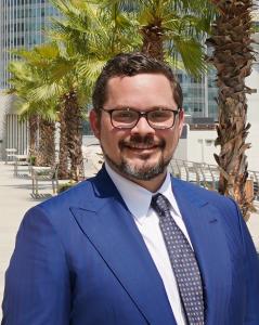 Johnny Kollin wearing a royal blue, pin-striped, suit, white shirt, purple tie with dots, and glasses, outdoors with palm trees and skyscrapers in the background.