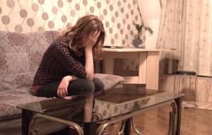 A woman sits on a sofa in front of a coffee table. She is resting face in her hand, her shoulder length light brown hair covers her face. Her posture gives the impression that she is upset.