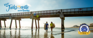 Visit Jacksonville photo of 2 surfers walking the beach by the pier with CAC badge and Visit Jax logo