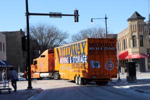 A large orange semi truck with the words "Midway Moving" written in navy lettering on the side turnes left in an intersection with two story historic Chicago brick buildings in the background.