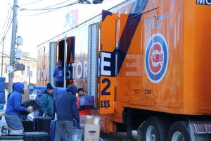 Movers wearing blue shirts and jackets transport items with the Chicago Cubs logo into a large orange semi truck with the words Midway Moving in navy lettering.