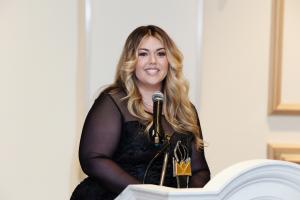 Brittany Bettini, Founder of I Need A VA. in black dress at podium speaking into a microphone