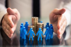 Syndication and Crowdfunding Concept. Figurines surrounding a stack of coins to demonstrate pooling funds into a project together. Protecting hands around the figurines and coins to demonstrate the syndicate/sponsor managing the deals.