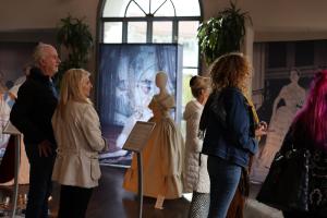 Visitors crowd around elegant dresses, holding glasses of wine, as they watch a depiction of Queen Elizabeth's coronation at the Couture Pattern Museum's exhibition.