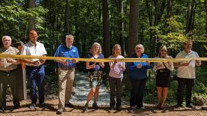 8 people, 3 men, 4 women, and another man, hold a ribbon and scissors. They are standing in front of a stand of trees on a mulched path with a new gravel path that leads to the new campsite behind them.