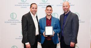 From left to right, IEDC President & CEO, Nathan Ohle, Grand Forks Region EDC Marketing & Communications Manager, Mark Maliskey, and IEDC Chair, Jonas Peterson stand together in front of a banner of the international economic development council logo.