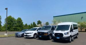 Four vehicles in a parking lot with a clear blue sky. The vehicles are a silver Honda Accord, a white BraunAbility Chrysler Voyager, a silver Chevy Express, and a white Transit Lift.