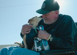 Man sitting in wheelchair outdoors with his small tan dog with a jacket on his lap. Their noses are touching.