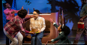 Production photo of a play - a young Black man sits on the back of a car holding a birthday cake and smiling while his family members gesture "Happy Birthday"