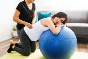 A pregnant woman leans over a stability ball while a doula massages her back.