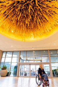  An indoor image featuring a woman in a manual wheelchair looking up at an impressive art installation on the ceiling. The installation radiates with warm yellow light and is composed of numerous wooden elements that resemble chairs, creating a striking g