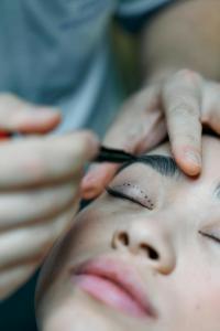 Close-up of a patient undergoing an eyebrow marking procedure in preparation for cosmetic surgery. The patient's closed eyelids show dotted lines indicating the areas to be addressed, while a practitioner's hands are carefully drawing the marks with a sur