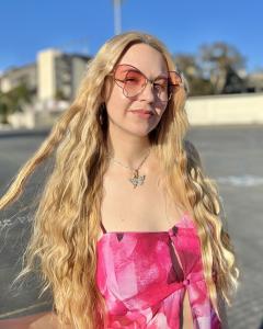 girl with long blonde wavy hair wears pink tank top, pink sunglasses, and a necklace with a butterfly pendant.