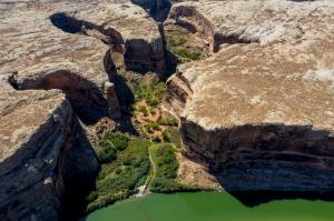 Ariel view of 4 white rafts parked along the side of the river leading up a side canyon, and sandstone walls are visible rising up to create the canyon. that is flush with green riparian vegetation. A trail is seen leading away from the river into the sid