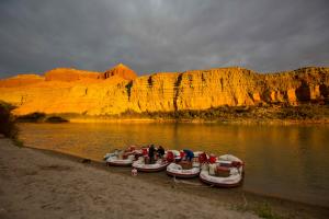 four white rafts are pulled up along a sandy beach on the side of the river. A couple guides are sitting on the raft or bending over. The evening sun creates a golden hue on the towering sandstone walls across the river.