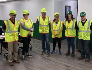 Professors in hard hats and safety vests pose for a photo before entering a massive metal building manufacturing plant.