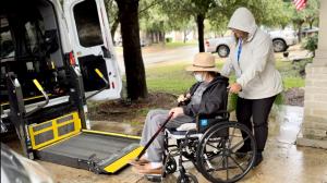 A female healthcare assistant and healthcare transport driver for Kerico company assisting a patient from his home and into a transport van for a trip to a medical appointment.