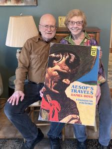 Man with Greek sailor's hat and pipe and woman with colorful blouse display poster for AESOP'S TRAVELS book