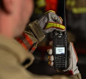 Photograph is looking over a firefighter's shoulder as they change the channel on the new Tait TP9900 portable radio. The officer is wearing heavy gloves.