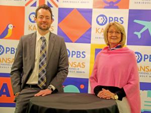 Brady Dreasher wearing a suit and Denice Morris wearing a pink shawl from Art of Estates stand in front of a colorful PBS banner with their hands on a round table