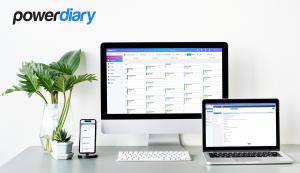 Power Diary practice management software on a desktop, laptop, and smartphone, showing a detailed appointment calendar. The setup includes a white desk with a keyboard and a vase with a monstera plant, creating a clean and professional workspace.