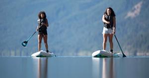 Two young girls on standup paddleboards each wearing life jackets and a leash in glassy, calm water on a beautiful sunny day with a mountainous terrain background