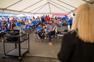 Dozens of people are seated, facing the camera, as they listen to CTCLUSI Lee Ann Wander speak, who is out of focus in the foreground.