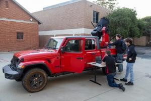 Three students working on a laptop connect to a hot red Jeep carrying an mobile optical station.