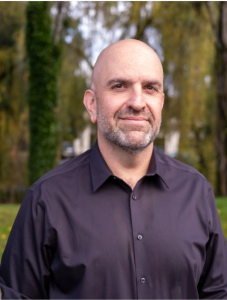 Amir wears a black shirt for this headshot photo outside in front of trees and grass