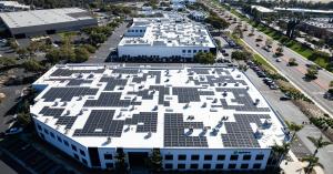 A drone shot of a large, two-story building with a larger solar system on the roof. The solar panel arrays are in various sizes to fit other things on the rooftop such as air conditioning units. There are other buildings in the background that also have s
