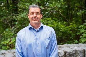 Lance Wells, Georgia Research Alliance Distinguished Investigator at UGA’s Complex Carbohydrate Research Center stands in front of a cement barrier with plenty of trees behind. He is smiling and is wearing a blue shirt.