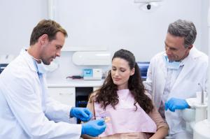 Two male dentists demonstrate a dental model to a female patient while explaining a procedure or treatment plan.