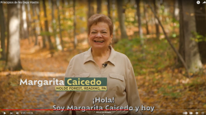 A screenshot captures a video clip of Margarita Caicedo smiling with her name captioned on the screen. It is a sunny fall day and her surroundings are of orange and yellow leaves on a trial at Nolde Forest Environmental Education Center.