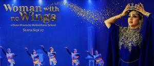Queen Cleopatra, dressed in royal blue affixes her crown to her head. Behind her, 5 dancers perform.