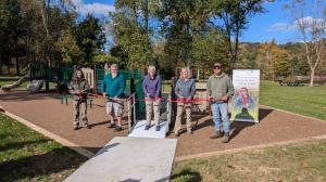 Five people spread out over 12 feet holding a ribbon in front of a new inclusive playground.