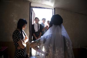 A young bride in a white wedding dress stands inside a room, with her hand on her hip, facing away from the camera and towards an open door to the outside, where the sunlight is bright. A group of three young women stand in the doorway, looking into the r