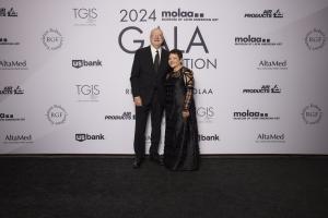 This image shows two individuals, a man and a woman, standing on a black carpet in front of a backdrop at an event. The backdrop displays the logos of various sponsors, including "molaa" (Museum of Latin American Art), "us bank," "AltaMed," and others. Th