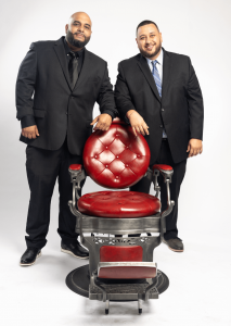 Luis Castillo and Tomas Castillo are standing on either side of a classic, vintage-style red leather barber chair at Miami Barber Institute. They are both wearing black suits, with the man on the left dressed in all black, and the man on the right wearing
