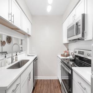 A kitchen with white cabinets, stainless steel appliances, and a sleek design. It features a sink, electric stovetop, microwave, and dishwasher. Light walls and dark flooring create a bright atmosphere, with a pass-through window offering a view of the living area.