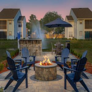 An outdoor seating area featuring a stone fire pit surrounded by blue chairs. Behind the fire pit is a bar with stools and an umbrella-covered table. In the background, there are apartment buildings and a fountain in the middle of a pond, creating a peaceful atmosphere.