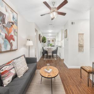 An open-concept apartment layout featuring a cozy living room with a gray sofa, abstract art, and hardwood floors. The space flows into a dining area and kitchen, with neutral walls, a ceiling fan, and modern décor, creating a comfortable and inviting atm