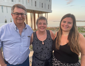 A photo of three people standing together outdoors in front of a modern building on a rocky shoreline. They are smiling, reflecting the warmth and unity that drives the Heather Cutler Foundation’s mission to improve pancreatic cancer care, particularly in