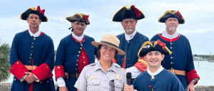 Group of people in historical costumes with tricorne hats along a waterfront.