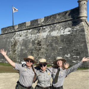 Three park rangers in uniform smile and pose with arms raised in front of a historic stone fort under a clear blue sky.