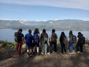 A group of Hood Canal Middle School students with backpacks stand on a hiking trail, looking out at the distant Olympic Mountains on a clear day. The students are engaged and connected with nature, representing teamwork and exploration.