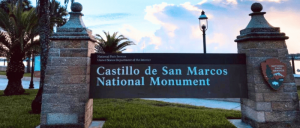 Sign for Castillo de San Marcos National Monument at dusk with lamppost and palm trees.