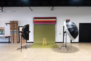 the studio has a white wall. in front of it is a green photo paper with a brown stool.