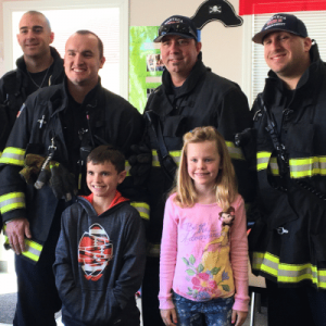 Three firefighters in gear stand with two children in a fire station.