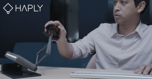 A person seated at a desk uses the MinVerse 3D mouse by Haply Robotics. The individual holds the device’s 2D mouse-like grip, while the MinVerse arm extends from its base on the desk into the air, allowing precise control in digital environments.