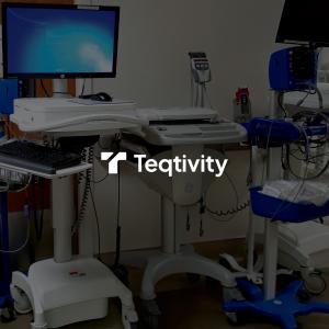 Medical room with multiple mobile computer workstations, including monitors, keyboards, and medical equipment on wheeled carts, with the Teqtivity logo overlaid on a darkened background. The workstations feature white adjustable stands and various connect